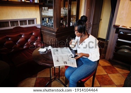 Stylish african american women in white blouse and blue jeans posed at cafe with newspaper and mobile phone, drink caramel latte. 