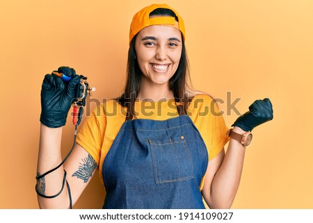 Young hispanic woman tattoo artist wearing professional uniform and gloves holding tattooer machine screaming proud, celebrating victory and success very excited with raised arm 