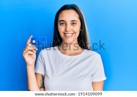 Young hispanic woman holding credit card looking positive and happy standing and smiling with a confident smile showing teeth 