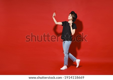 Full length side view of funny young brunette woman 20s in basic black t-shirt hat stand doing selfie shot on mobile phone showing victory sign isolated on bright red color background studio portrait