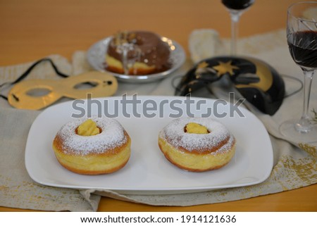 Wooden table with carnival food: home baked donut with homemade vanilla cream; in background donuts with chocolate coating, two glasses of red wine and carnival masks