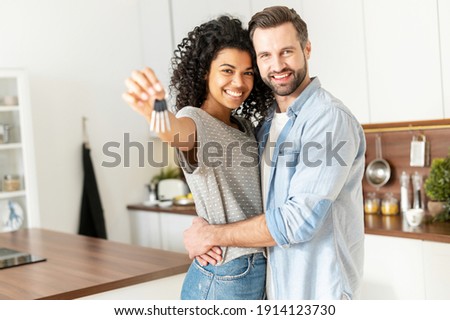 Excited multiracial couple in love move in a new apartment. Cheerful African woman in embrace of a caucasian guy holds the keys, the newlywed look at the camera and smile Royalty-Free Stock Photo #1914123730