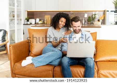 Cheerful diverse couple in love spends leisure time online with a laptop at home. Young woman and man look at the laptop screen and laugh sitting on the sofa, watching comedy movies, funny videos Royalty-Free Stock Photo #1914123925