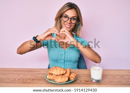 Beautiful caucasian woman eating croissant for breakfast smiling in love doing heart symbol shape with hands. romantic concept. 