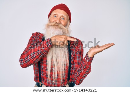 Old senior man with grey hair and long beard wearing hipster look with wool cap amazed and smiling to the camera while presenting with hand and pointing with finger. 