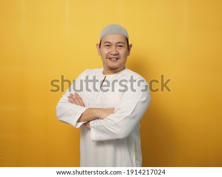 Portrait of Asian muslim man smiling friendly with crossed arms, confident gesture, successful businessman, against yellow background
