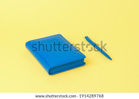 A blue notebook and a blue ballpoint pen on a yellow background. Monochrome image of office accessories.