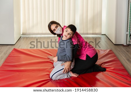 disabled child and physiotherapist on a red gymnastic mat doing exercises. pandemic mask protection