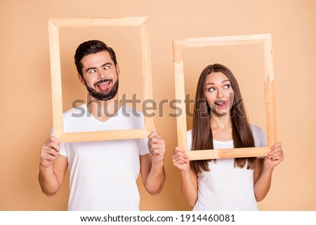 Photo of sweet childish husband wife wear white t-shirts fooling holding wooden frames isolated beige color background