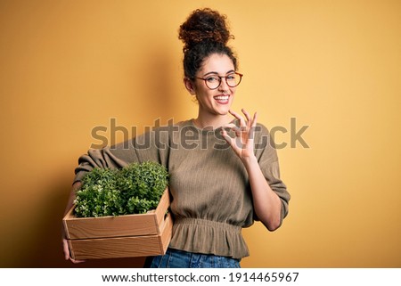 Young beautiful gardener woman with curly hair and piercing holding wooden box with plants doing ok sign with fingers, excellent symbol