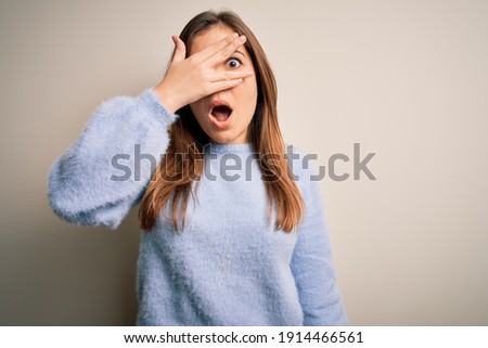 Beautiful young woman wearing casual winter sweater standing over isolated background peeking in shock covering face and eyes with hand, looking through fingers with embarrassed expression.