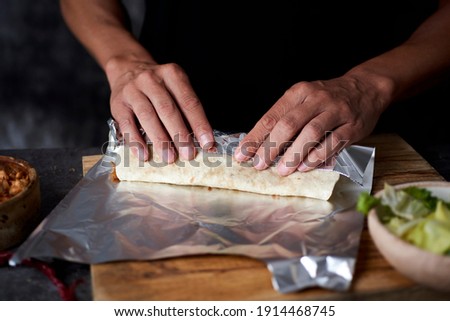 a young man at a table wraps in aluminum foil a durum or a burrito, filled with chicken meat cooked with different vegetables such as onion or red and green pepper Royalty-Free Stock Photo #1914468745