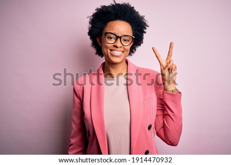 Young beautiful African American afro businesswoman with curly hair wearing pink jacket smiling looking to the camera showing fingers doing victory sign. Number two.