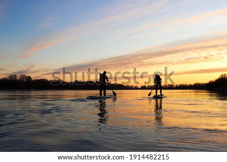 Silhouettes of two people on SUP (stand up paddle board) during a colorful sunset in the calm winter Danube river