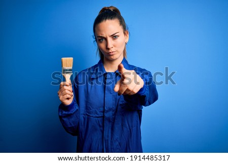 Young beautiful painter woman with blue eyes painting wearing uniform using paint brush pointing with finger to the camera and to you, hand sign, positive and confident gesture from the front