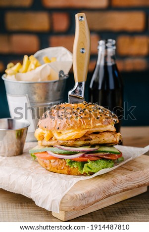 Delicious juicy burger from Brioche Bun. Chicken steak, cheese, onion, tomato, cucumber, lettuce, stuck knife, sauce, drink and fries. Cafe table. Wooden board