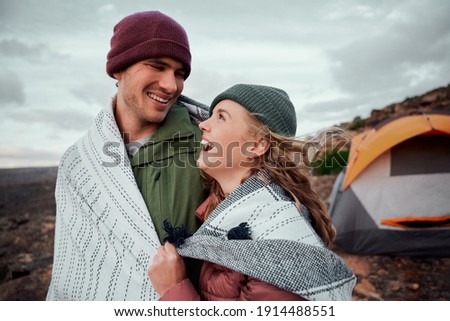 Laughing young romantic couple in a blanket standing together outside tent on mountain looking at each other