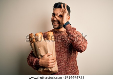 Young handsome man holding groceries paper bag of fresh baguette bread over isolated background with happy face smiling doing ok sign with hand on eye looking through fingers
