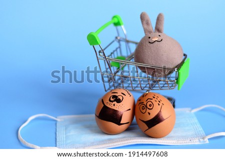 Creative Easter eggs and toy rabbit with Corona virus (COVID19) protection concepts. Chicken eggs on mini shopping cart with doodle faces wearing medical masks on blue background. Flat lay, top view. 