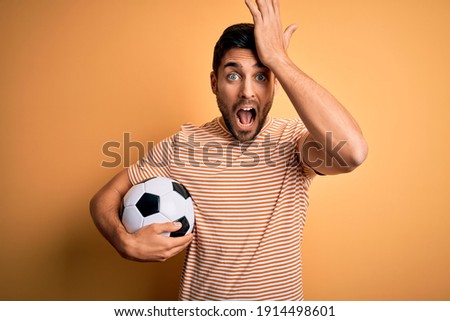Handsome player man with beard playing soccer holding footballl ball over yellow background surprised with hand on head for mistake, remember error. Forgot, bad memory concept.