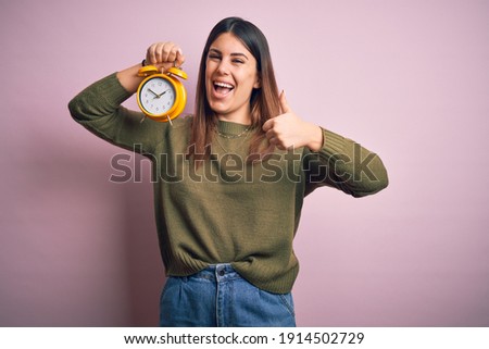 Young beautiful woman holding alarm clock standing over isolated pink background happy with big smile doing ok sign, thumb up with fingers, excellent sign