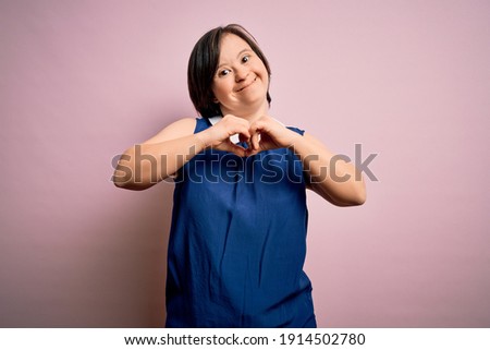 Young down syndrome woman wearing elegant shirt over pink background smiling in love showing heart symbol and shape with hands. Romantic concept.