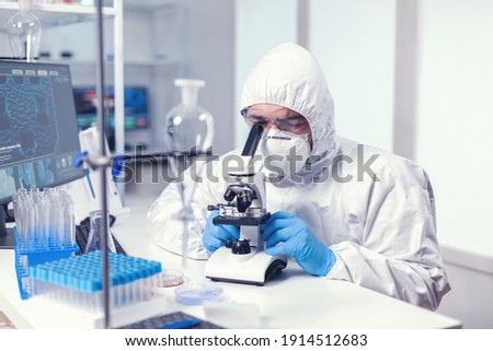 Analyzing virus in microbiology lab using microscope wearing ppe suit and glasses. Virolog in coverall during coronavirus outbreak conducting healthcare scientific analysis. Royalty-Free Stock Photo #1914512683