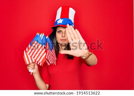 Young beautiful brunette patriotic woman wearing united states hat holding usa flags with open hand doing stop sign with serious and confident expression, defense gesture
