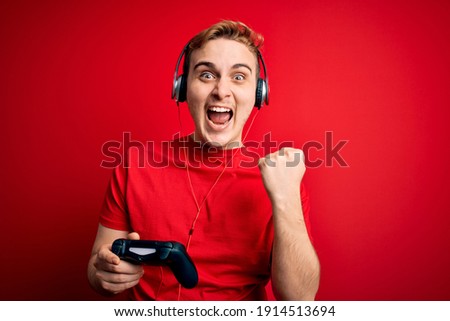 Young handsome redhead gamer man playing video game using headphones and joystick screaming proud, celebrating victory and success very excited with raised arms