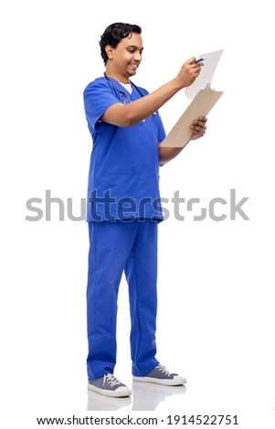 healthcare, profession and medicine concept - happy smiling indian doctor or male nurse in blue uniform writing medical report on clipboard over white background
