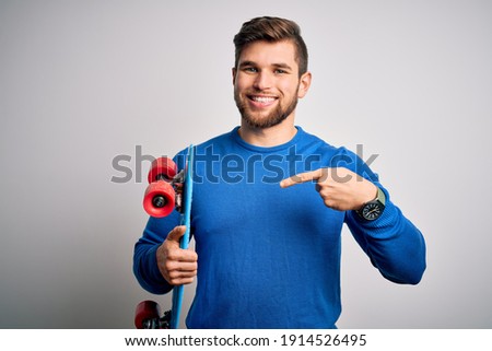 Young blond skeater man with beard and blue eyes holding skate over white background very happy pointing with hand and finger