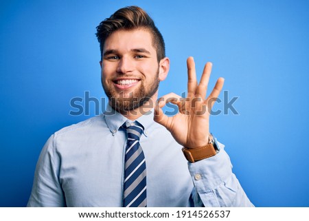 Young blond businessman with beard and blue eyes wearing elegant shirt and tie standing smiling positive doing ok sign with hand and fingers. Successful expression.