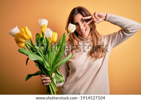 Young beautiful brunette woman holding bouquet of yellow tulips over isolated background Doing peace symbol with fingers over face, smiling cheerful showing victory