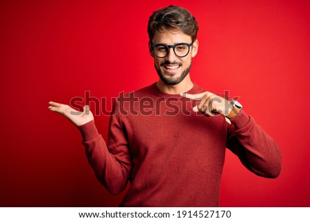 Young handsome man with beard wearing glasses and sweater standing over red background amazed and smiling to the camera while presenting with hand and pointing with finger.