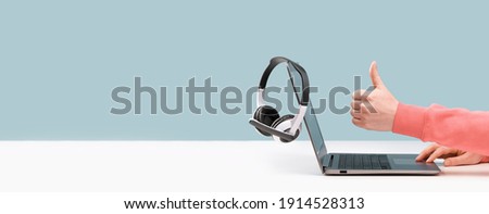 Side view of slim laptop and like thumb up hand. wireless headphones on grey desk. Blue background. Distant learning. working from home, online courses support. Audio podcast. vlogger blogger banner Royalty-Free Stock Photo #1914528313