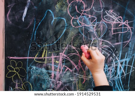 Little girl child draws with the right hand close-up pink chalk on a black wooden board, easel picture, doodle. Home creativity. Drawing lesson at school.