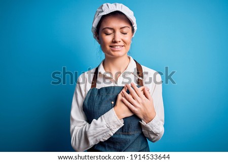 Young beautiful baker woman with blue eyes wearing apron and cap over blue background smiling with hands on chest with closed eyes and grateful gesture on face. Health concept.