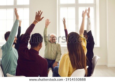 Group of happy diverse young and mature people in casual clothes sitting on chairs in circle and raising hands, approving of good idea and voting for useful suggestion in community club meeting Royalty-Free Stock Photo #1914550888