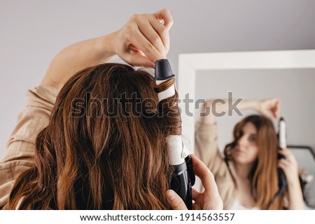 Young woman styling her shiny long brown hair with curling iron, looking at mirror at home. Selective focus, low depth of field Royalty-Free Stock Photo #1914563557