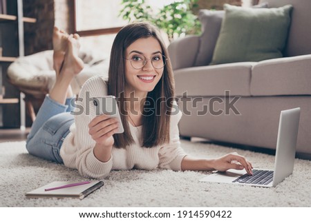 Full size photo of beautiful joyful woman hold phone use laptop lie floor charming smile inside home indoors house