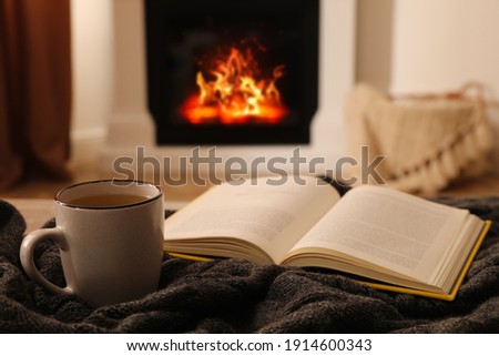 Cup of hot tea and book on knitted plaid near fireplace at home. Cozy atmosphere Royalty-Free Stock Photo #1914600343