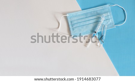 Medical mask, syringe and ampoule on a two-tone white and blue background with copy space. The concept of vaccination, treatment and immunity enhancement.