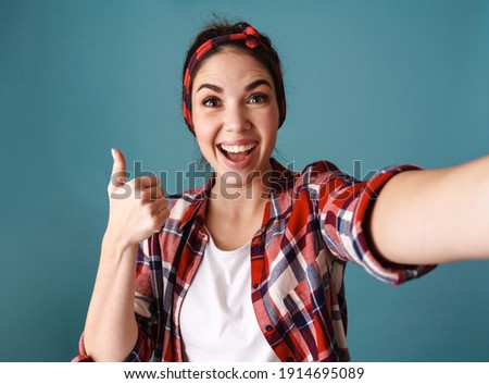 Joyful young woman showing thumb up while taking selfie photo isolated over blue background