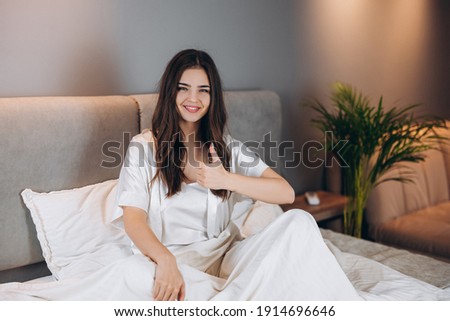 female model in white silk pajamas shows cool. Young beautiful woman with a smile on her face sitting on the bed shows okay