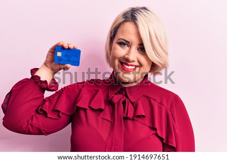 Young beautiful blonde plus size woman holding credit card over isolated pink background looking positive and happy standing and smiling with a confident smile showing teeth