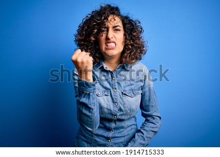 Young beautiful curly arab woman wearing casual denim shirt standing over blue background angry and mad raising fist frustrated and furious while shouting with anger. Rage and aggressive concept. Royalty-Free Stock Photo #1914715333
