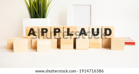 Wooden cubes with letters on a white table. The word is APPLAUD. White background with photo frame, house plant.