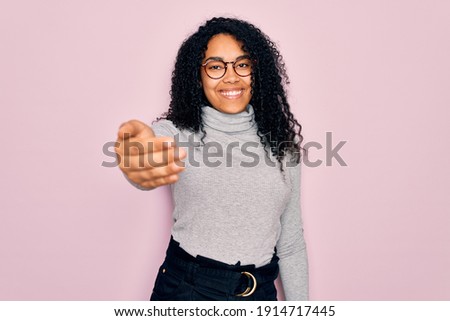 Young african american woman wearing turtleneck sweater and glasses over pink background smiling friendly offering handshake as greeting and welcoming. Successful business.
