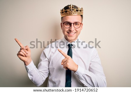 Young handsome caucasian business man wearing golden crown over isolated background smiling and looking at the camera pointing with two hands and fingers to the side.