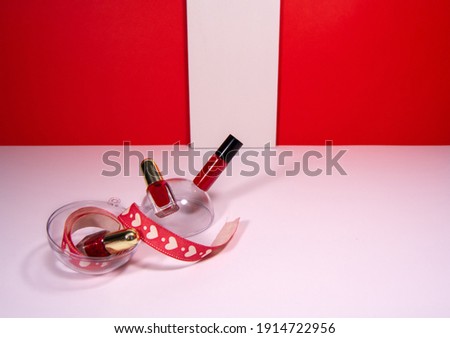 Valentine's Day background. Red lipstick and nail polish on white table with white stripe on red background. Ribbon with hearts lies in a round shape. Copy space for you text